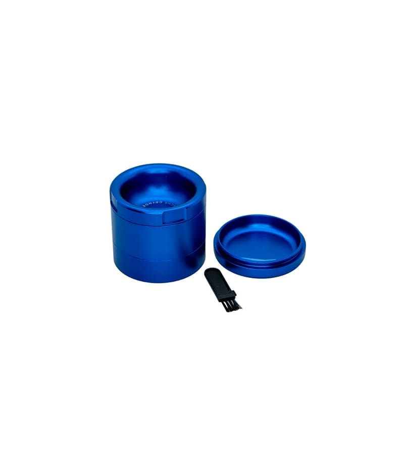 A blue metal grinder with its top removed, revealing "GRINDER" text inside, next to a small black brush.