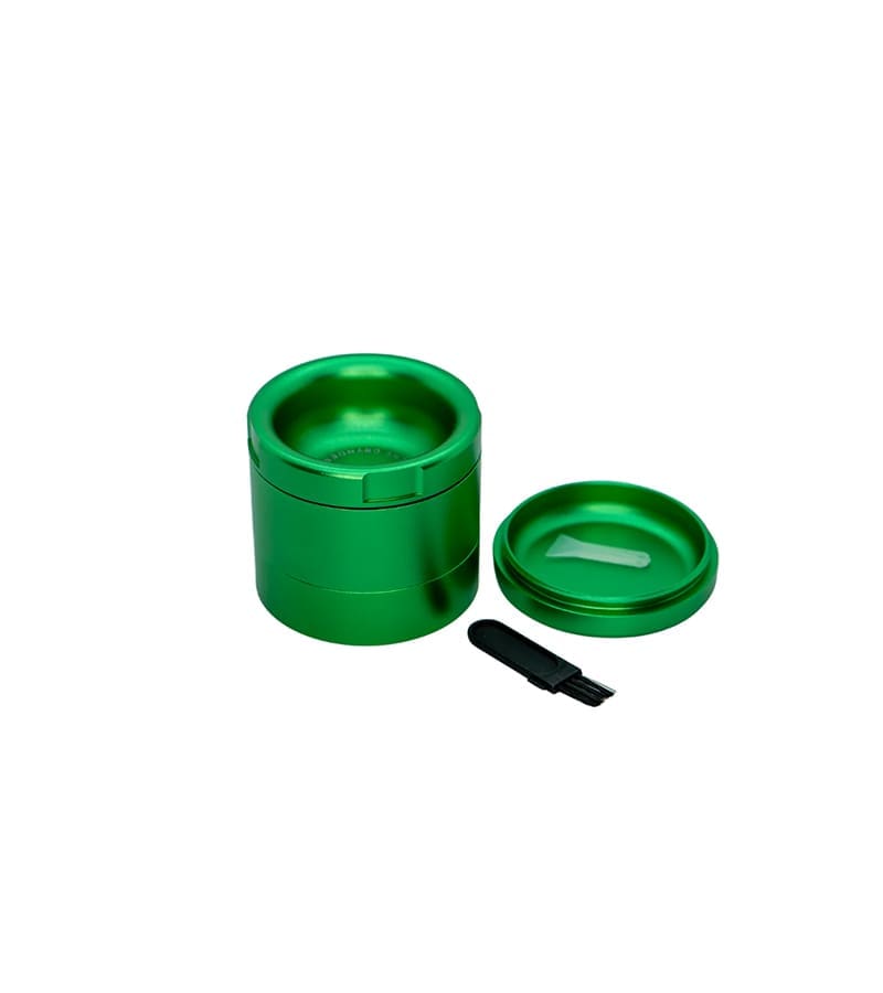 A shiny green herb grinder with its lid off, a small white scraper, and a black brush on a white background.