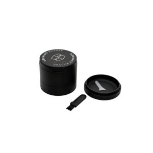 A black metal grinder with "STACHE GRYNDER" text and logo, a lid, and a small brush.
