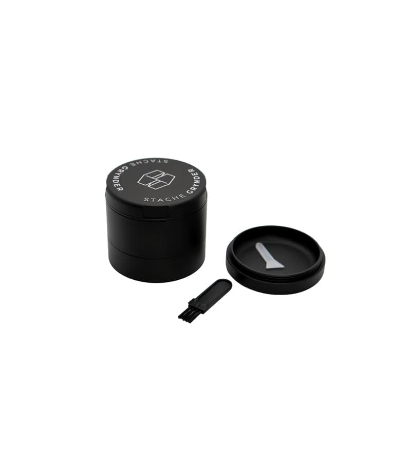 A black metal grinder with "STACHE GRYNDER" text and logo, a lid, and a small brush.