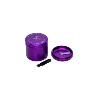 A purple "STACHE GRYNDER" with its lid off, showing a white scoop inside, and a small black brush nearby.