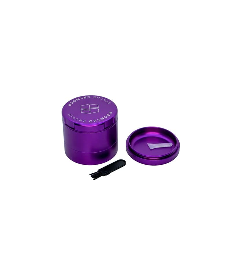 A purple "STACHE GRYNDER" with its lid off, showing a white scoop inside, and a small black brush nearby.