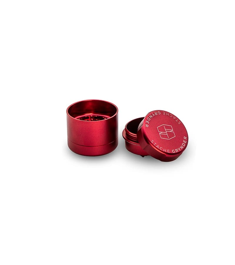 A red metal grinder with the lid slightly open, revealing "STACHE GRYNDER" and a logo.